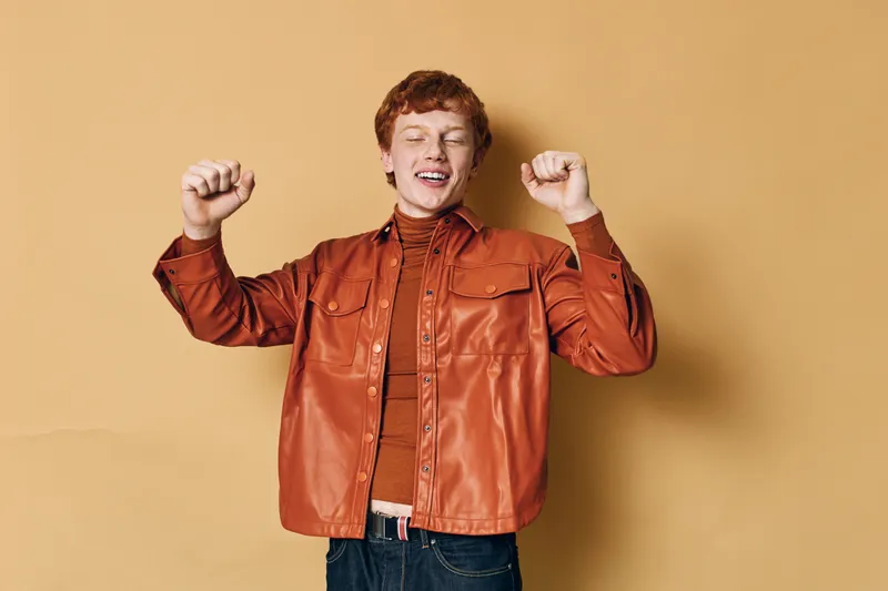 Young man with red hair wearing brown leather jacket stands with raised fists and closed eyes expressing excitement or celebration against beige background.