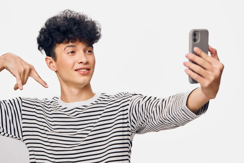 young man taking selfie with smartphone, wearing striped shirt, smiling and showing hand gesture on white background, casual lifestyle moment