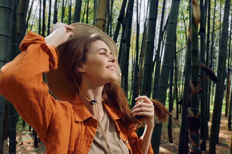 Woman in bamboo forest wearing a hat and orange jacket, smiling in nature. Travel portrait of young woman enjoying sunlight and green trees, casual outfit and peaceful mood outdoors.