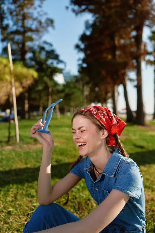 Smiling woman in modern boho-western denim street style with red bandana enjoys sunny outdoor lifestyle with film color tones and natural green background.