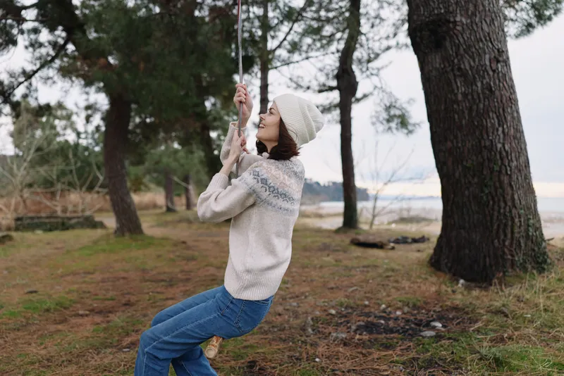 Woman on a swing outdoors among trees by a lakeside, wearing knit sweater and beanie, enjoying a calm moment in nature and fresh air.
