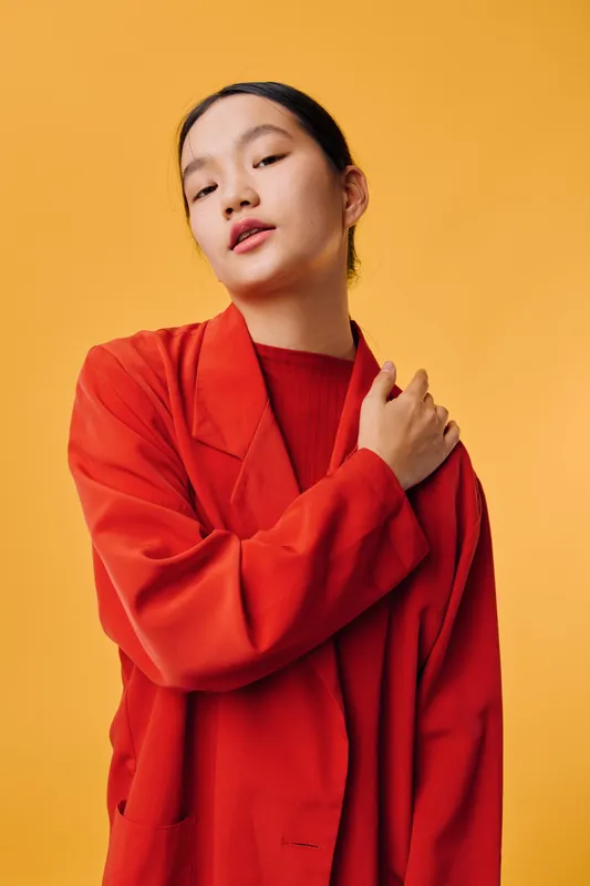 Young woman posing in oversized red coat with hand on shoulder against orange background, fashion portrait with confident expression and casual style in studio lighting.
