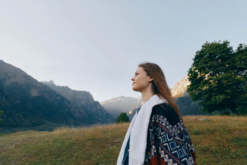 Woman portrait in mountains nature meadow outdoors landscape travel scene, long hair and patterned coat, serene profile facing horizon, peaceful mood, fresh air and golden light.
