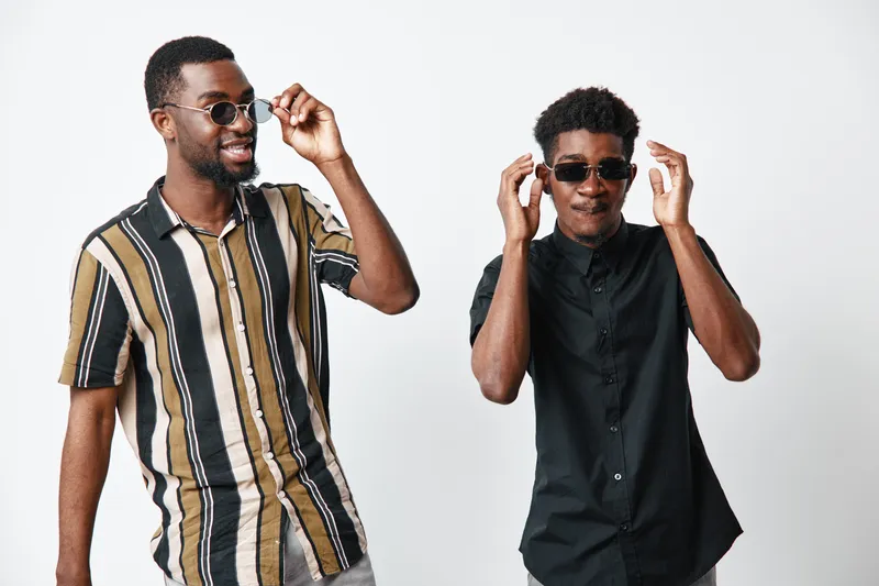 Two young black men with dark skin wearing sunglasses and casual shirts posing and smiling on white background for lifestyle and friendship concept.