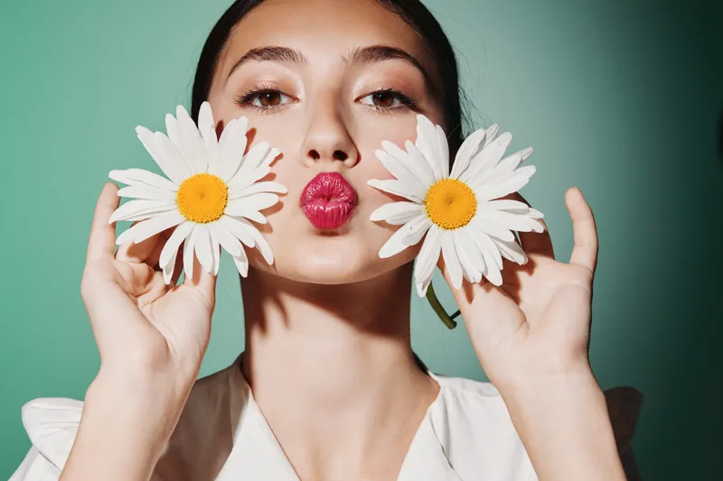 Young woman holding daisies near face with bright lipstick posing on green background. Concept of beauty, fashion, creative, fresh, and playful portrait with natural flowers.