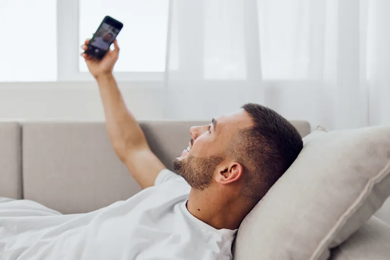 Smiling man using smartphone while relaxing on a couch in a bright, modern living room. Perfect for lifestyle and technology themes in 2025 and beyond.