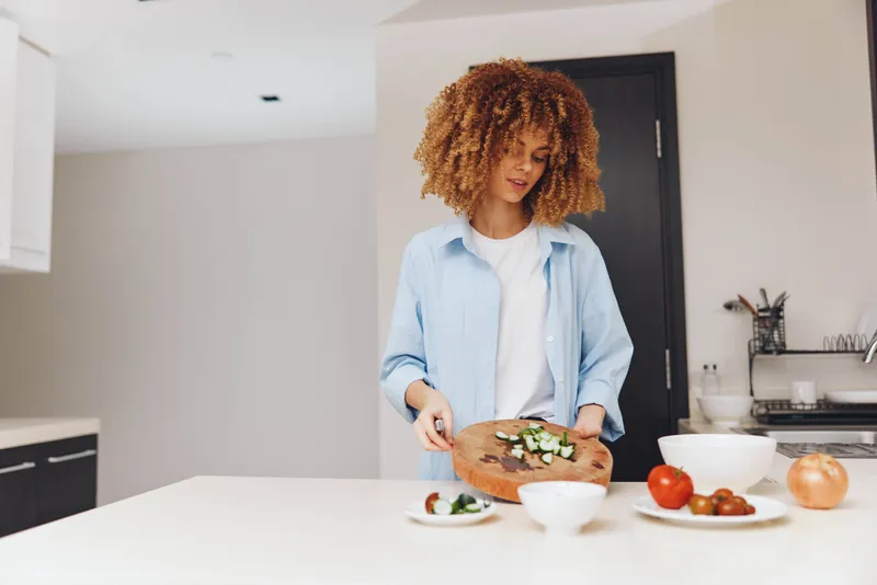 Женщина с вьющимися волосами стоит на кухне, держа миску с едой на стойке, в уютной домашней обстановке