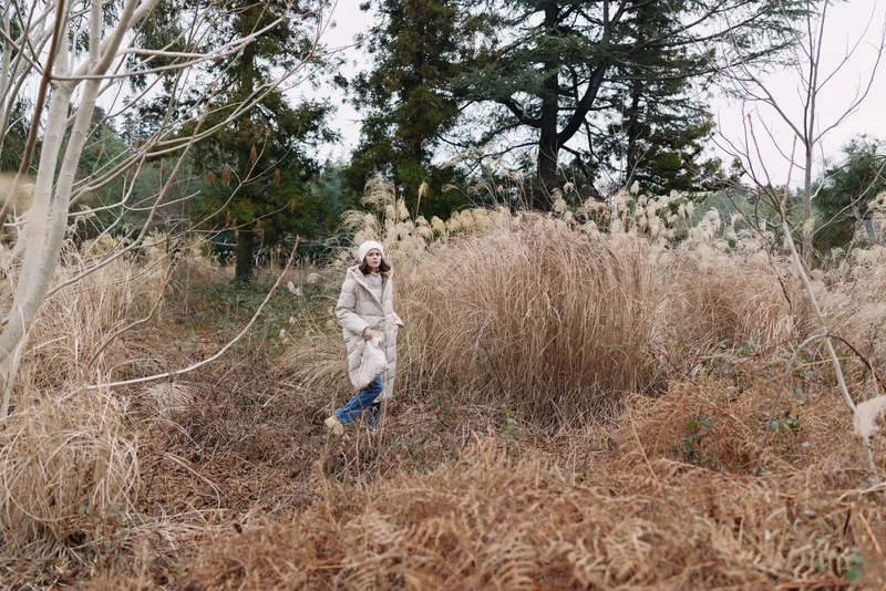 Person walking through a field of tall dried grass in winter, wearing a light coat, surrounded by bare trees and muted landscape, conveying solitude and calm