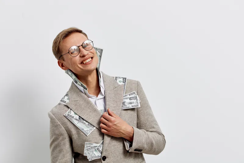Smiling woman with glasses holding money and with dollar bills on her beige blazer, cheerful expression, internal studio portrait, business and finance concept, success, happiness, wealth, professiona
