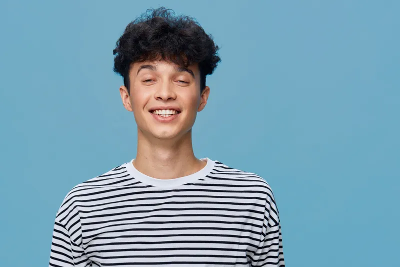 smiling young man with curly hair wearing striped shirt, expressing happiness on blue background in casual style, natural lighting, cheerful mood