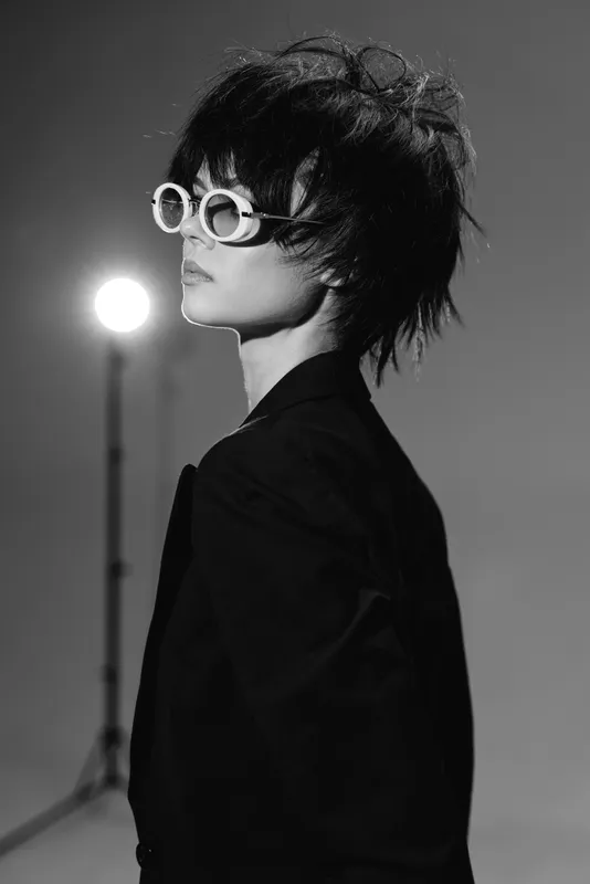 Black and white portrait of a stylish woman wearing sunglasses and a black blazer with a modern hairstyle in studio lighting.