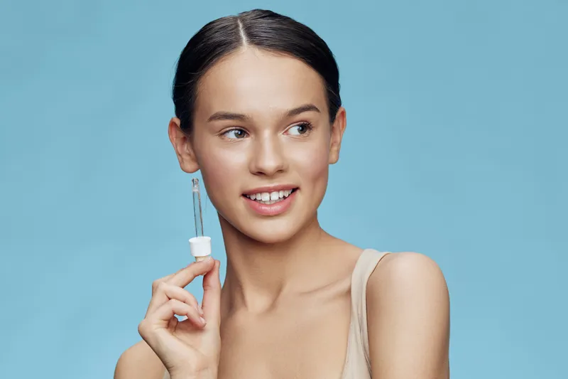 Beautiful young woman with glowing skin holding a dropper bottle in front of a blue background, representing skincare and beauty concepts