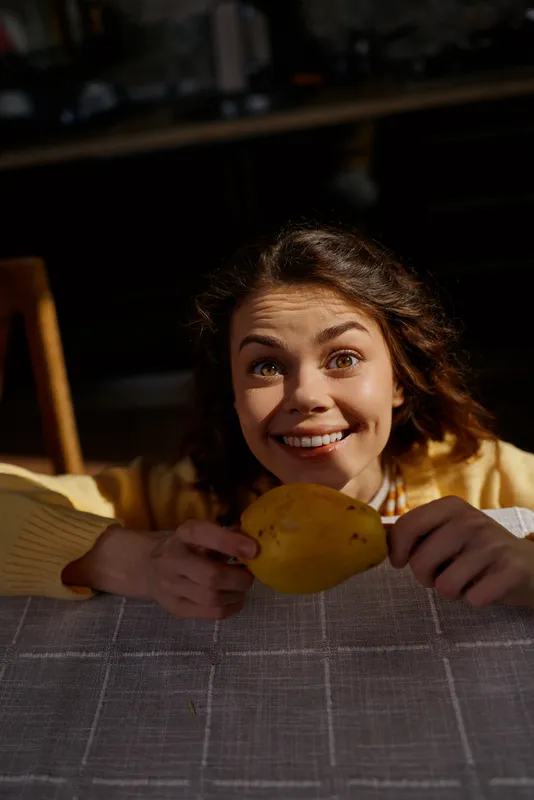A cheerful woman leans over a table holding a bright lemon, smiling warmly at the camera in a cozy home setting, casual lifestyle moment, fresh fruit scene, captured during a relaxed morning, soft