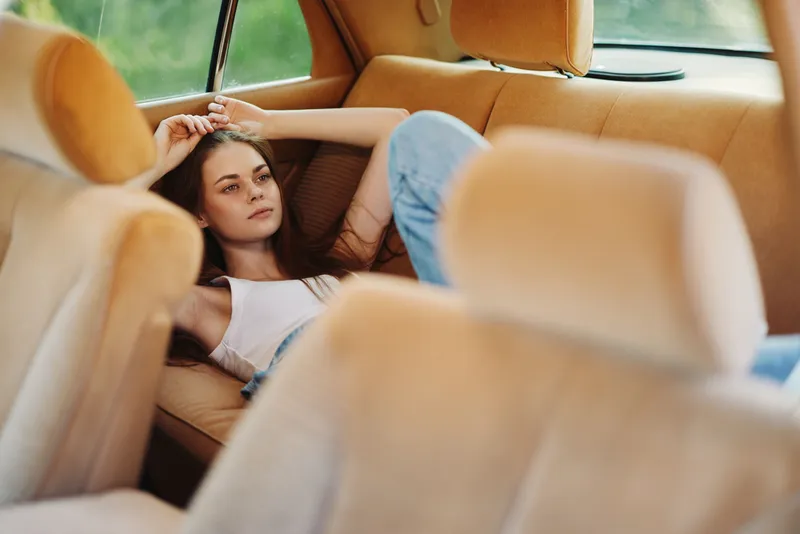 Young woman relaxing in a vintage car interior, wearing casual attire, expressing serenity and comfort, with a soft-focus green background highlighting tranquility.