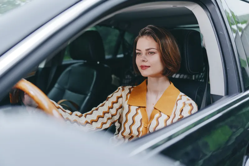 A woman sits behind the steering wheel with a gentle smile, poised in a car interior ready for a journey.