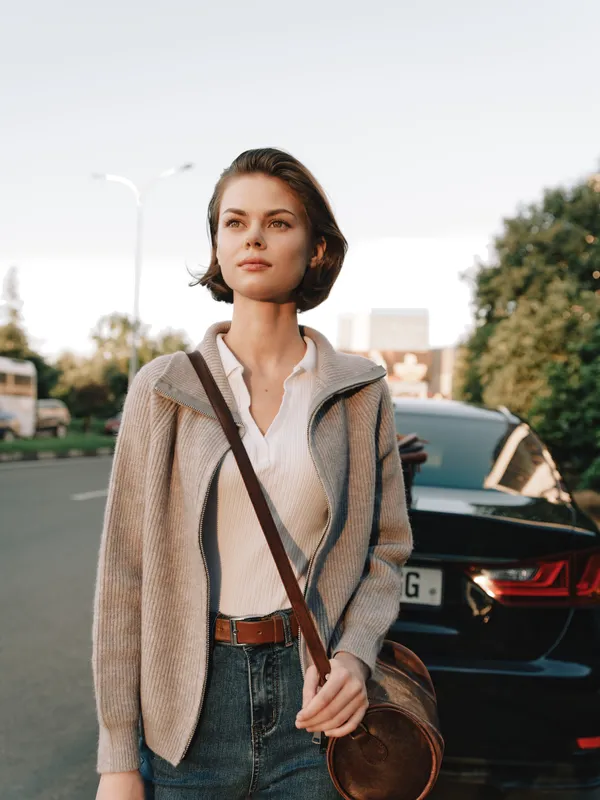 A stylish woman stands on a city street, looking into the distance. She wears a beige cardigan over a white shirt and denim jeans, with a crossbody bag, exuding calm urban confidence.