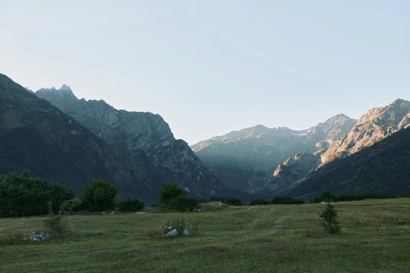 A serene mountain valley is captured in soft natural light. The rugged peaks rise against a clear sky, with a lush meadow and scattered trees creating a peaceful landscape.