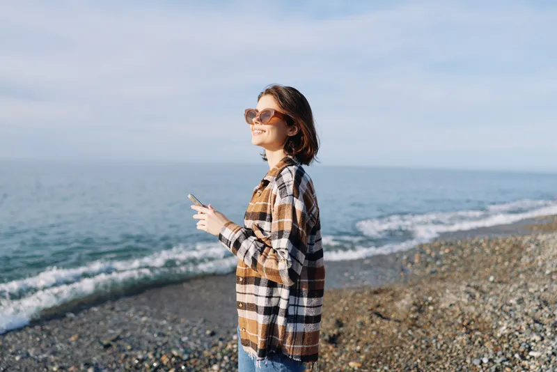 Woman on a beach in a plaid shirt and sunglasses, casual outdoors style by the sea, relaxed portrait at the coastline, sunlit ocean scene