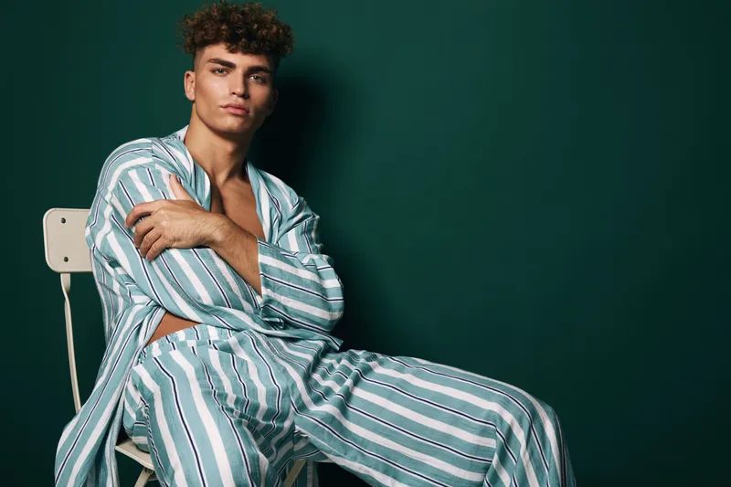 Young man with curly hair wearing striped pajamas sitting on chair with arms crossed against dark green background in studio. Casual fashion, relaxed pose and confident expression.