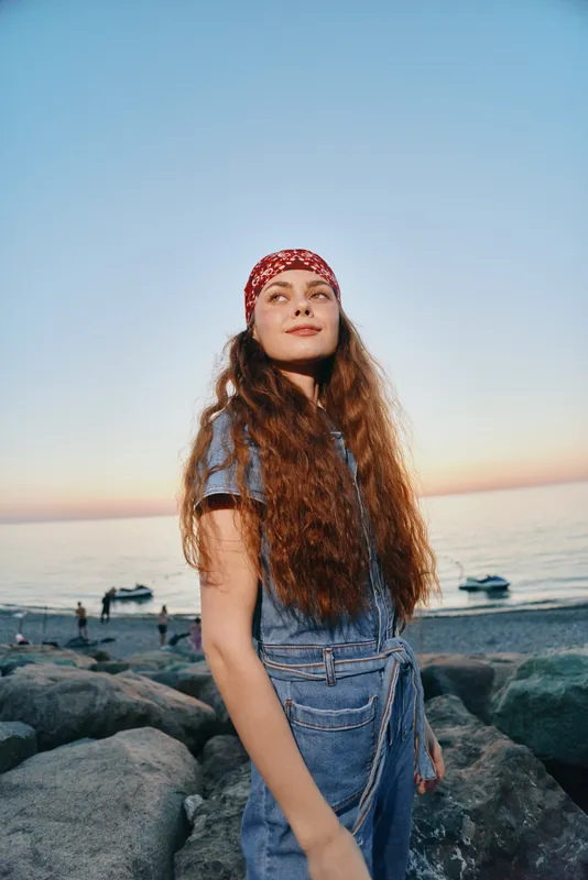 young woman with long hair wearing denim dress and red headscarf standing on rocky beach at sunset with calm sea background