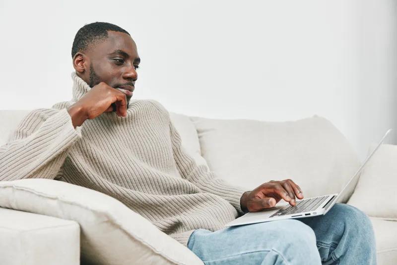 Thoughtful young man working on a laptop from home, wearing a cozy sweater in a modern living room setting showcasing a relaxed atmosphere