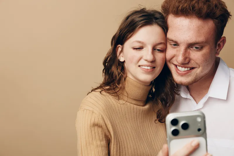 Young couple with happy expressions looking at smartphone screen together. Casual clothing and warm beige background create cozy atmosphere. Concept of technology and relationship.