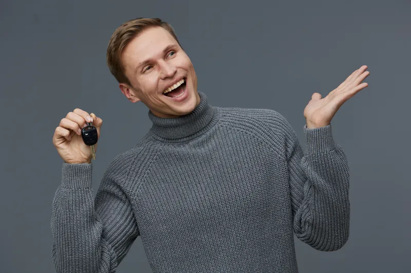 Smiling man with short brown hair wearing a cozy gray turtleneck sweater, joyful expression, holding a car key, gesturing happily with one hand, isolated on plain neutral background, studio portrait. 