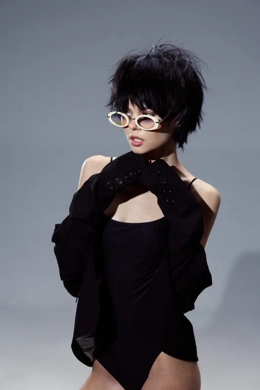 Stylish woman in black outfit and white sunglasses posing on gray background with confident expression