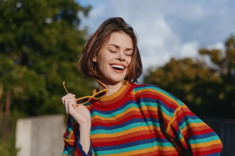 Young woman in her early 20s wearing a colorful striped sweater smiling and laughing outdoors, holding yellow sunglasses; candid lifestyle portrait, autumn fashion casual outfit in park sunlight.