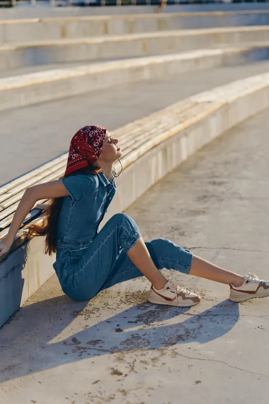 young woman in denim jumpsuit sitting outdoors on concrete steps, wearing red headscarf and white sneakers, enjoying sunny warm day, casual style