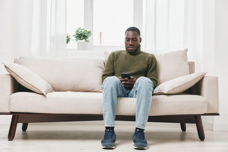 Thoughtful Black man sitting on a light sofa, checking his phone, in a bright and modern living room filled with natural light and greenery