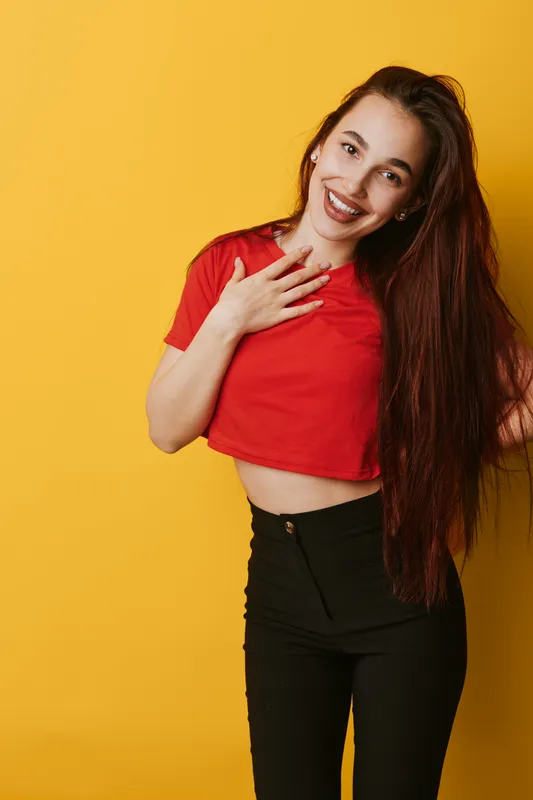 Young woman with long hair smiling and touching chest in casual red top and black pants against yellow background. Portrait of happy female with natural makeup and earrings.