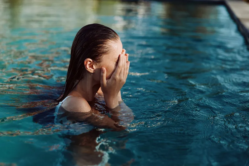 Woman in a pool covers her face with her hands, water glistening around her. A moment of relaxation, wellness, and self care in a warm, sunlit outdoor setting.