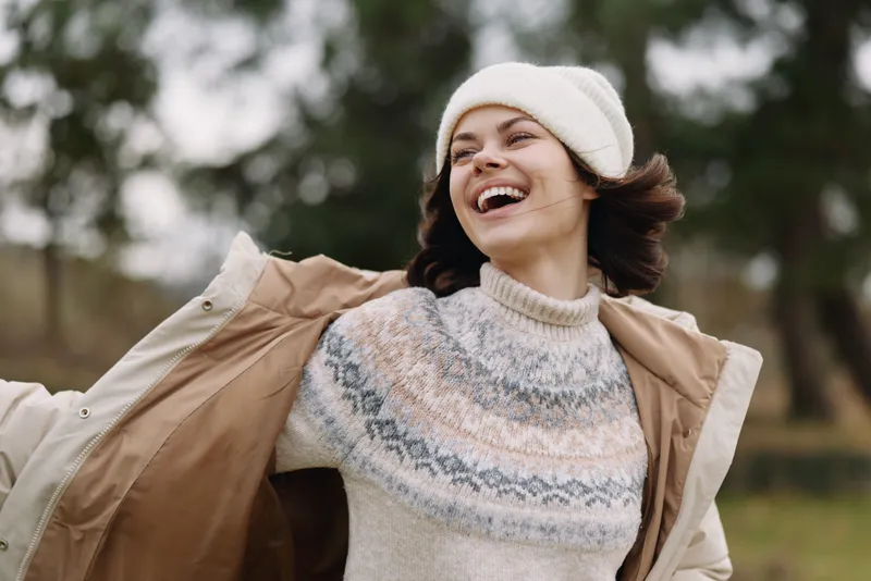 Joyful woman outdoors, laughing with arms outstretched, wearing a knit sweater, beanie and light coat, in a windy park setting, capturing a carefree moment and seasonal style against vivid autumn