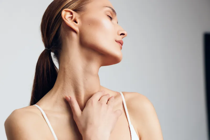 Young brunette woman in a white top turns her head to the side, showing a graceful profile with a slender neck, soft studio lighting, and a serene, introspective mood.