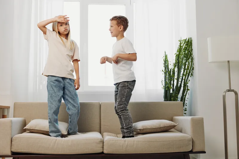 Two children standing on a beige sofa in a bright living room with natural light, casual clothes, and a tall green plant near a modern lamp. Concept of childhood energy and play.