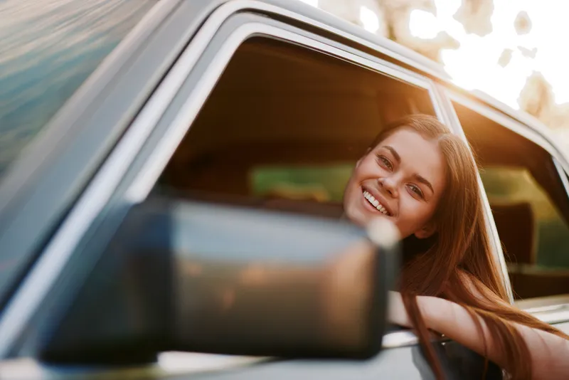 Happy young woman leaning out of a car window with long hair, enjoying a sunny day, conveying joy and freedom in a vibrant outdoor setting.