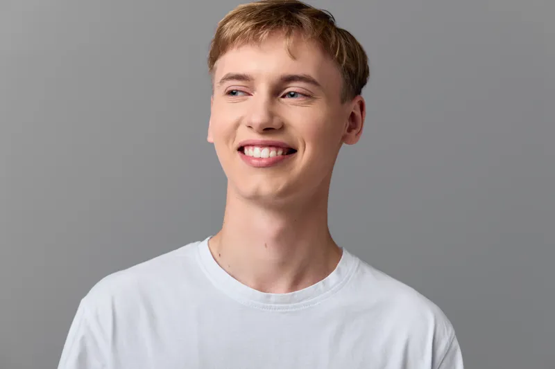 Smiling, young man wearing a white shirt in a studio with a neutral gray background, showcasing confidence, approachability and modern, clean style for lifestyle and headshot use.