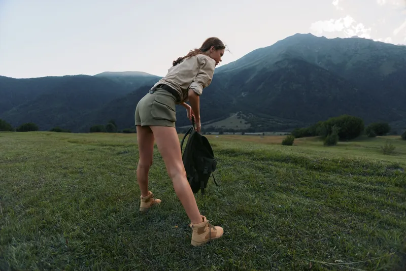 Young woman hiking across a lush meadow toward distant mountains, carrying a jacket and backpack, enjoying a breezy outdoor moment amid wide green scenery.
