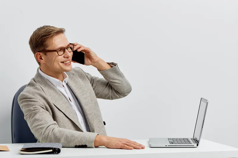 Professional man talking on mobile phone while sitting at desk with laptop isolated on plain background. Business lifestyle, communication, workplace, technology, modern office. Studio shot in color.