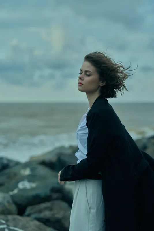 Windy seaside scene with a calm figure wearing a dress and dark coat, standing on rocks near the ocean, hair blowing, moody sky, serene moment of contemplation and solitude by the shore.