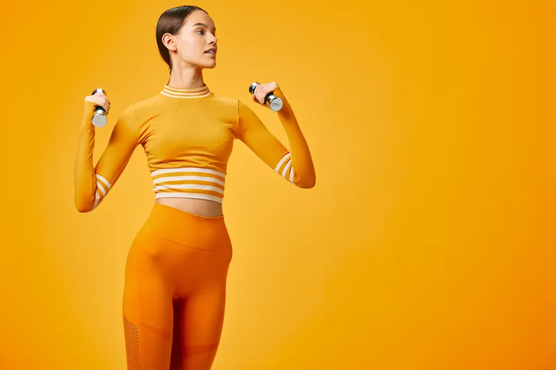 Woman fitness and sport theme: fit young woman in matching orange activewear holding light dumbbells, performing a shoulder exercise in a minimal studio with a vibrant orange background.