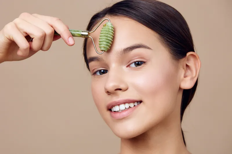 Young woman performing facial massage with a green jade roller, showcasing a glowing complexion and a smile against a soft beige background, promoting skincare wellness