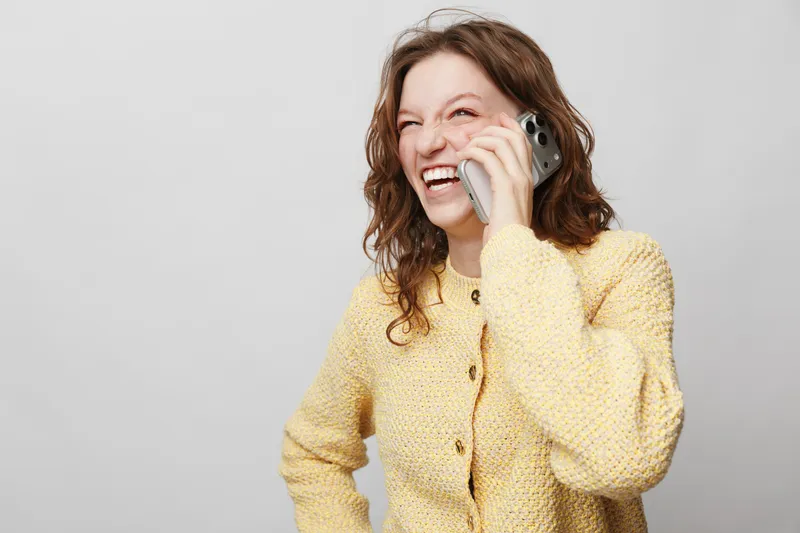 Happy woman with curly hair wearing yellow sweater laughs while talking on smartphone. Isolated on light background. Concept of communication, joy, casual lifestyle, and positive emotions.