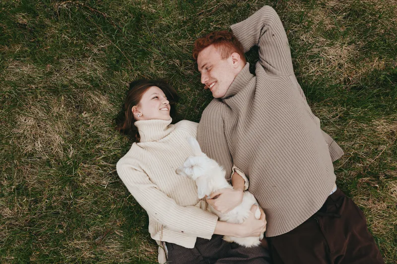 A couple in cozy sweaters lounge on a grassy field, smiling at each other while holding a small white dog. A warm, candid moment captures casual happiness, connection, and relaxed outdoor lifestyle.
