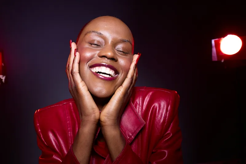 Happy African woman with dark skin smiles joyfully, hands on face, wearing red leather jacket. Studio lighting creates vibrant mood on dark background. Concept of beauty and confidence.