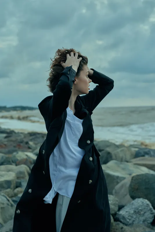 Woman in a dark coat battles wind on a rocky coast, white shirt fluttering, capturing moody outdoor fashion by the sea. Windy day drama.
