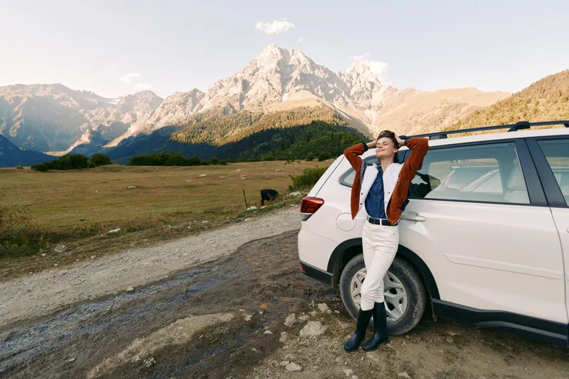 Mountain woman leans against car at roadside in open landscape, enjoying travel and adventure in a scenic field. Gravel road, snowcapped peaks, warm jacket, boots and relaxed exploration.