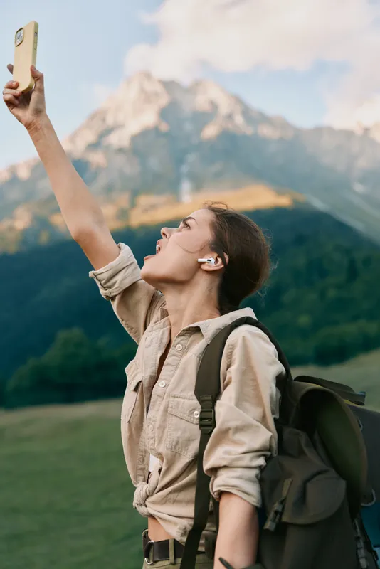 Young woman outdoors raises her phone for a selfie as alpine peaks rise behind her, capturing a spontaneous moment of adventure, confidence, and joy.