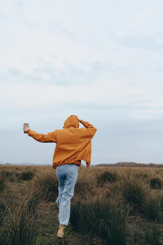 Woman in orange jacket standing in outdoor field, arms out, back view, windy nature under cloudy sky, lifestyle freedom mood, casual travel feeling in open air, for personal growth and peace